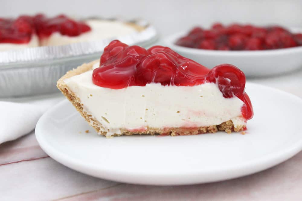 slice of cream cheese pie topped with cherry pie filling on white plate on marble counter with pie in background