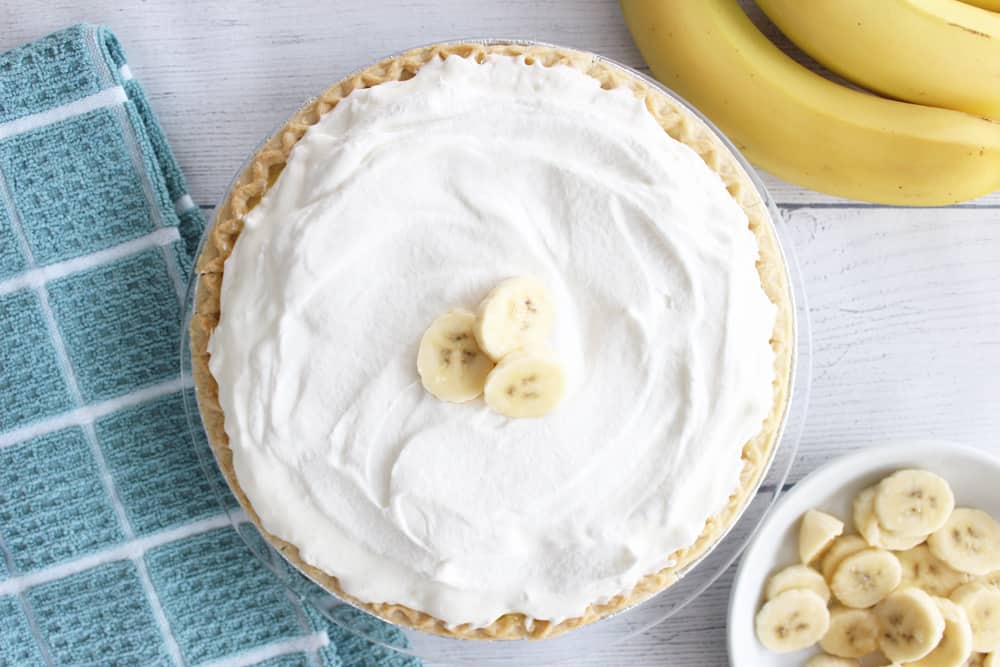 a full banana cream pie next to a blue towel, 3 bananas and some sliced banana on a white plate