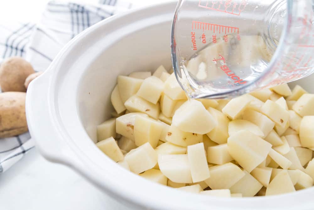 Peeled and chopped potatoes in a slow cooker with water being poured into the bowl.
