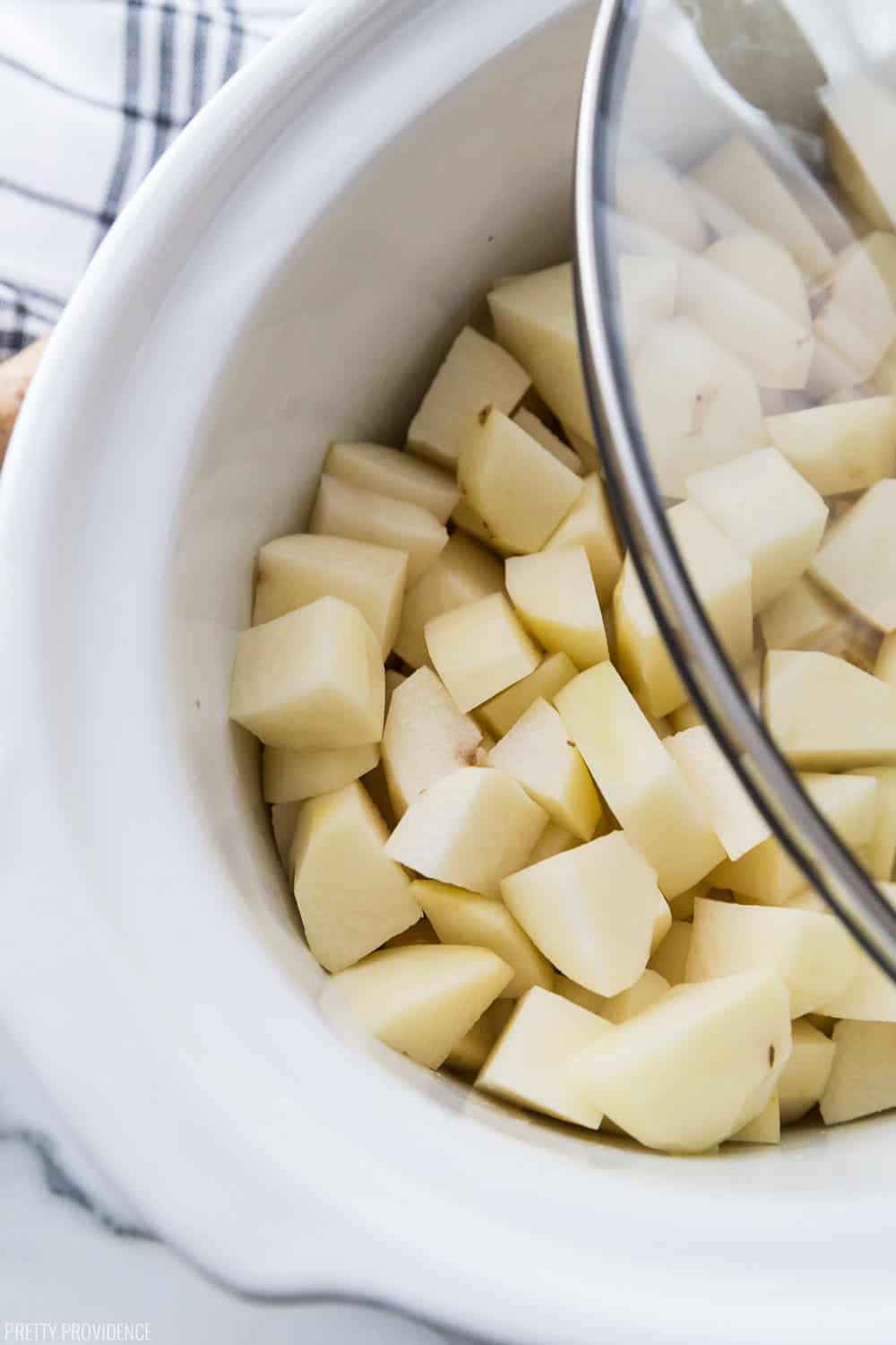 Peeled and chopped potatoes in a white slow cooker with lid being put over it.