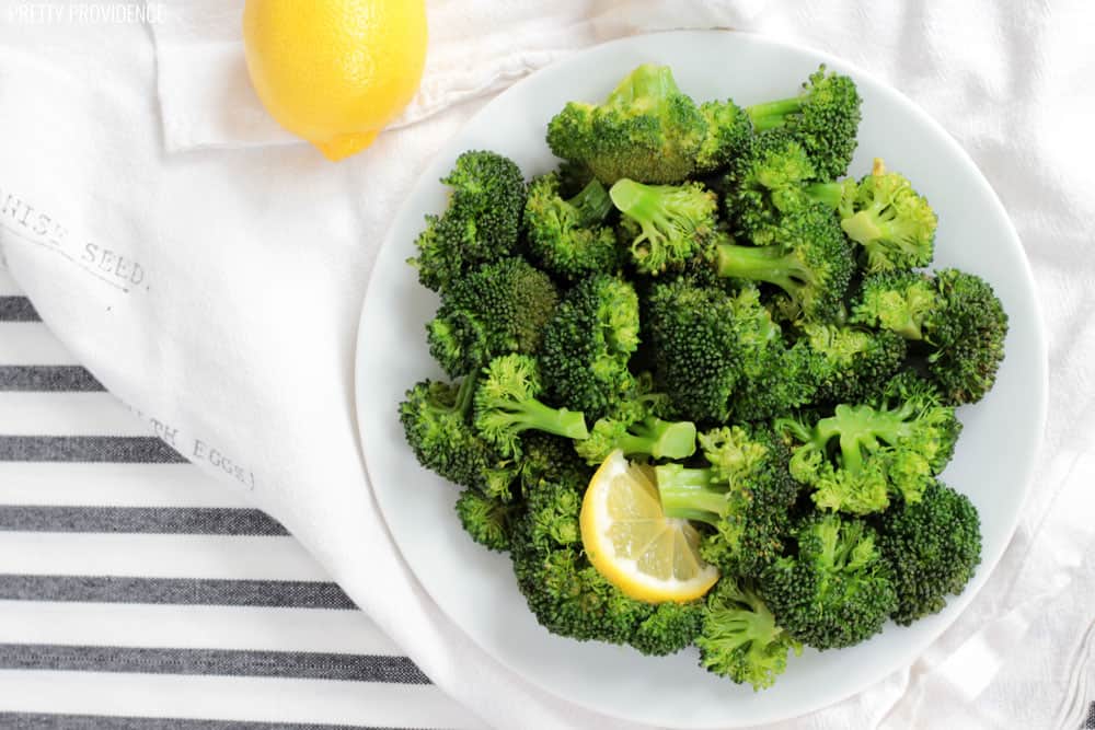 a plate full of steamed broccoli on a white linen tea towel with a lemon for garnish
