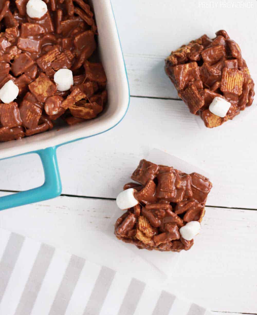 Two squares of Golden Grahams S'mores Bars on a white wood table with the pan of treats next to them.