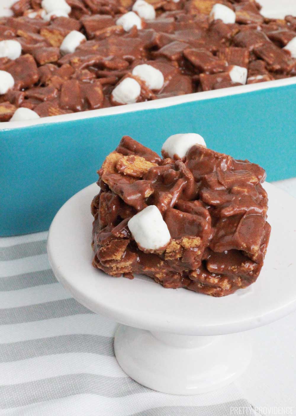 Golden Grahams S'more Bar square with marshmallows on a small cake stand in front of a pan of s'mores treats.