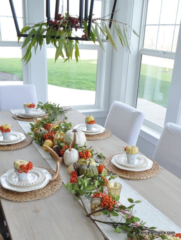 Thanksgiving Tablescape Orange, white, green pumpkins, greenery and berries on a burlap table runner.