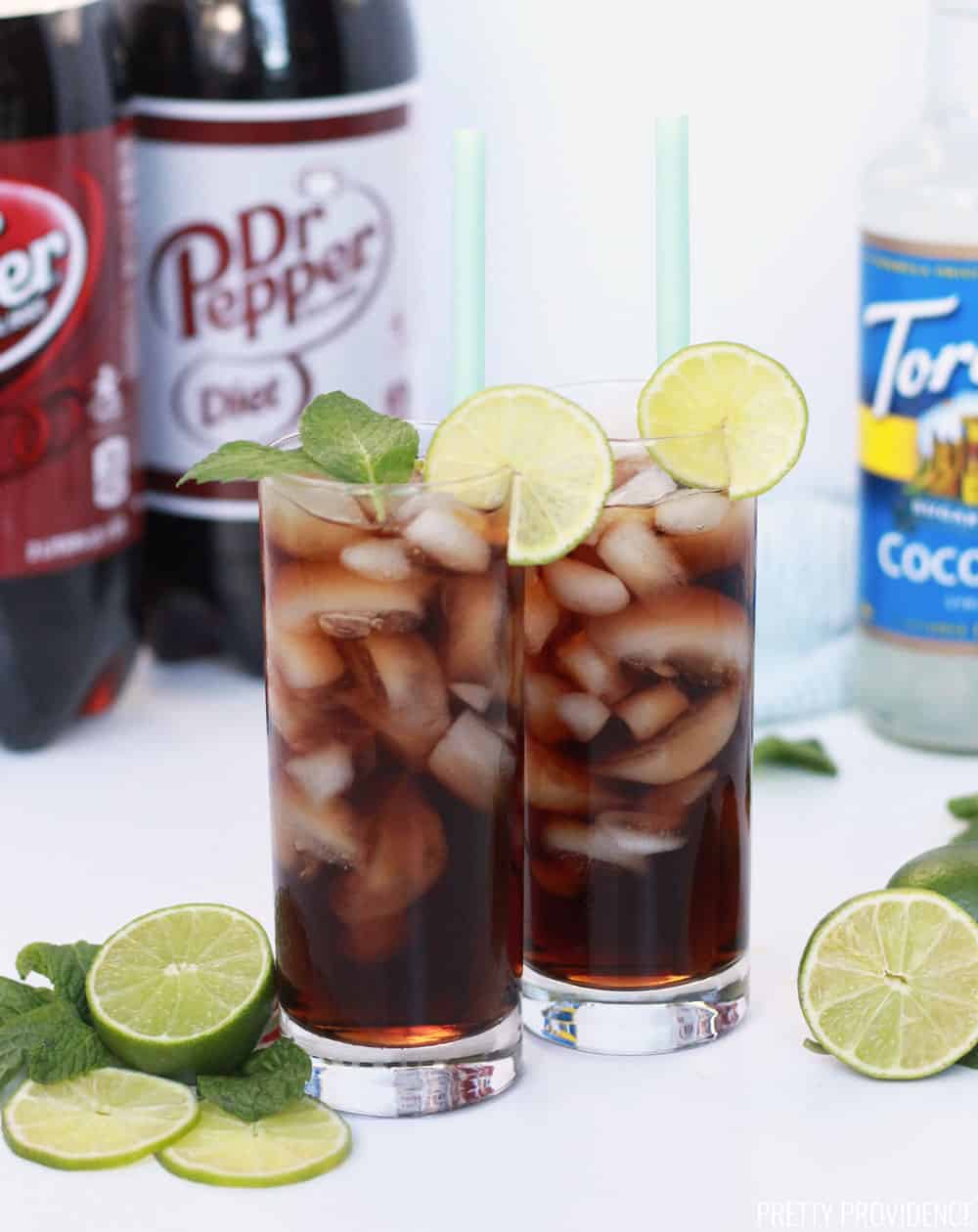 Dirty Dr. Pepper drinks with Dr. Pepper bottles and Coconut syrup in the background, lime slices and mint as garnish.