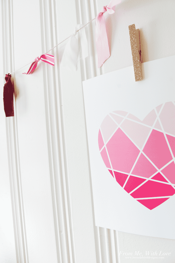 Geometric heart with light pink sections on top, medium pink in the middle and dark pink at the bottom, hung up on a white wall with a clothes pin and a Valentine's Day garland.