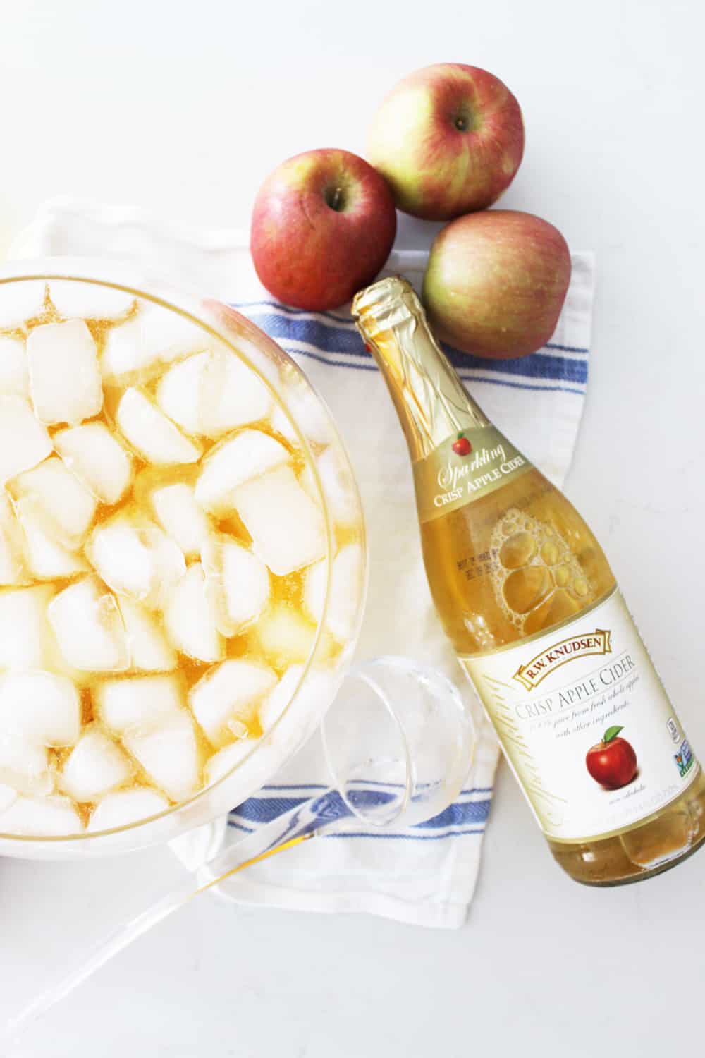 a punch bowl of apple cider with ice next to three apples and a bottle of cider