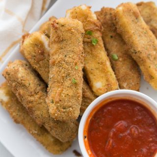 a stack of homemade cheese sticks on a white platter next to a small white dish of marinara sauce