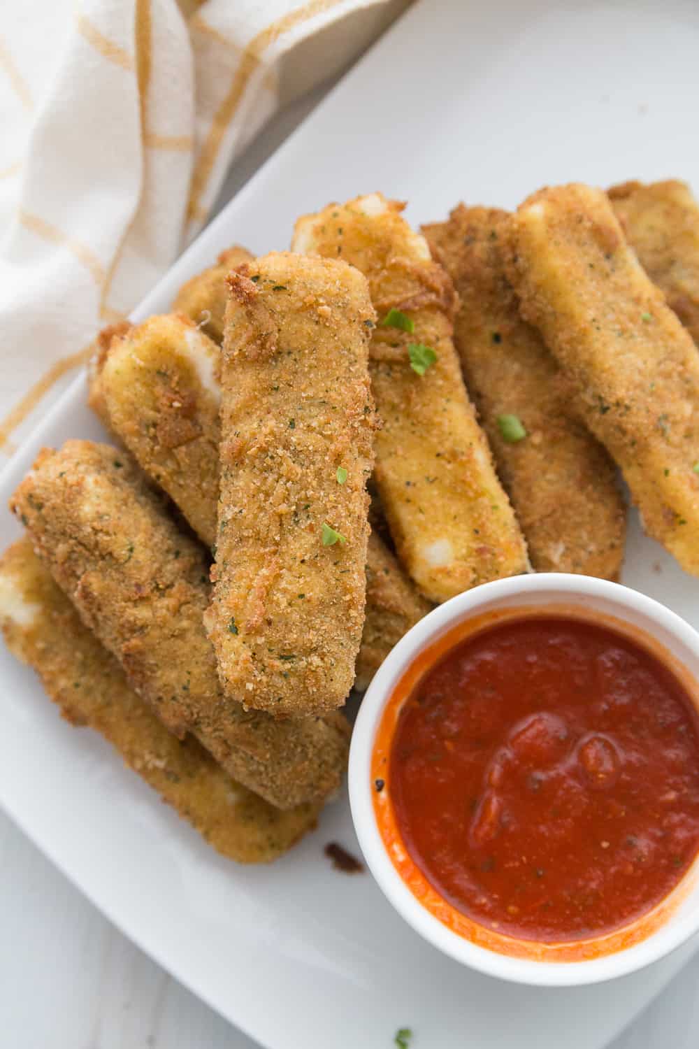 a batch of homemade mozzarella sticks on a white platter next to a small ramekin of marinara sauce