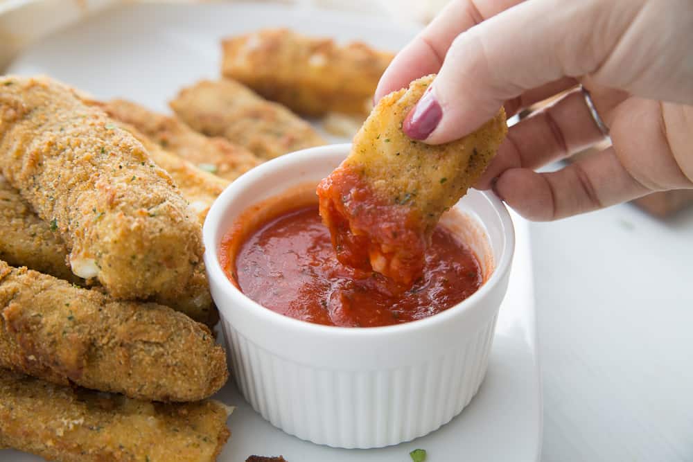 a half eaten mozzarella stick being dipped in marinara sauce in a white ramekin