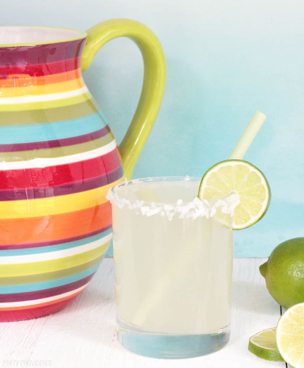 Limeade in a glass with coconut shavings on the rim. A colorful pitcher is in the background.