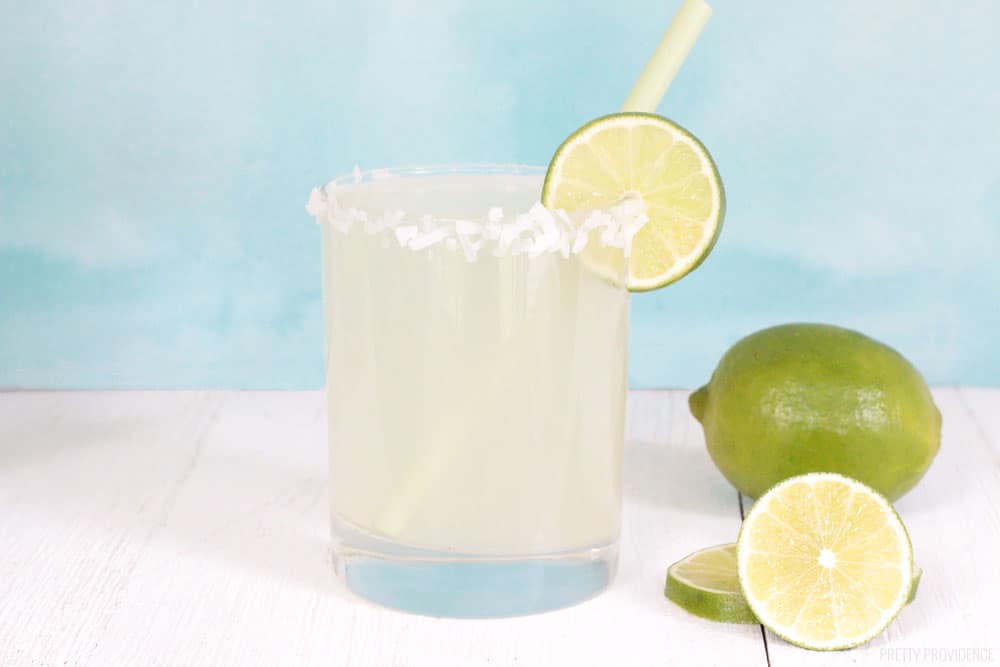 Coconut Limeade in a glass with shredded coconut and a lime slice on the rim, and sliced limes on the side.