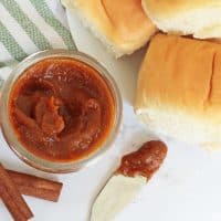 Pumpkin Butter in a small jar, two cinnamon sticks, a knife with pumpkin butter on it and freshly baked rolls.