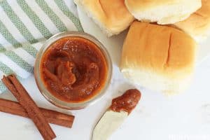 Pumpkin Butter in a small jar, two cinnamon sticks, a knife with pumpkin butter on it and freshly baked rolls.