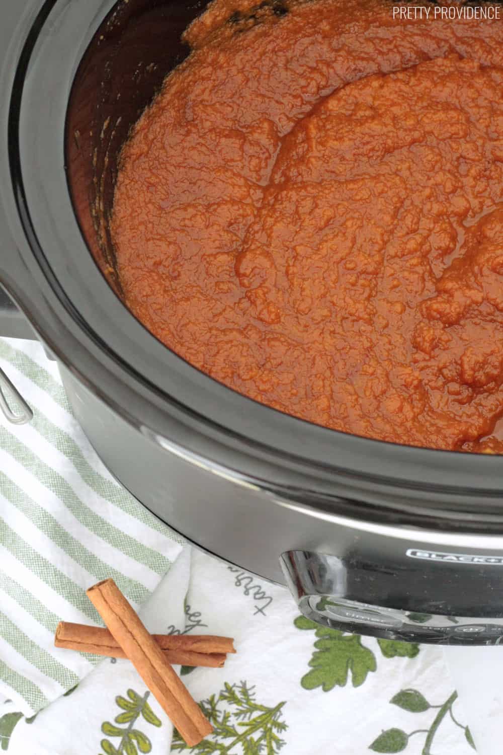 Pumpkin Butter in a slow cooker with cinnamon sticks and fall tea towels next to the slow cooker.