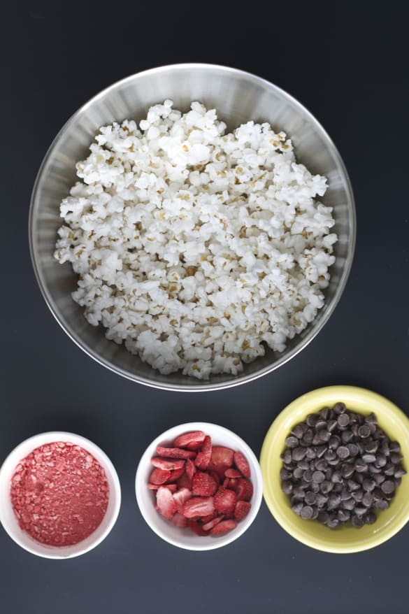 Large bowl of popcorn, small white bowls with freeze dried strawberries and dark chocolate chips on a black surface.