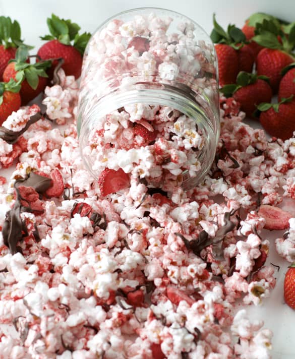 Chocolate Covered Strawberry Popcorn - Pink popcorn with freeze-dried strawberries and dark chocolate drizzled on top, spilling out of a glass jar with strawberries in the background.