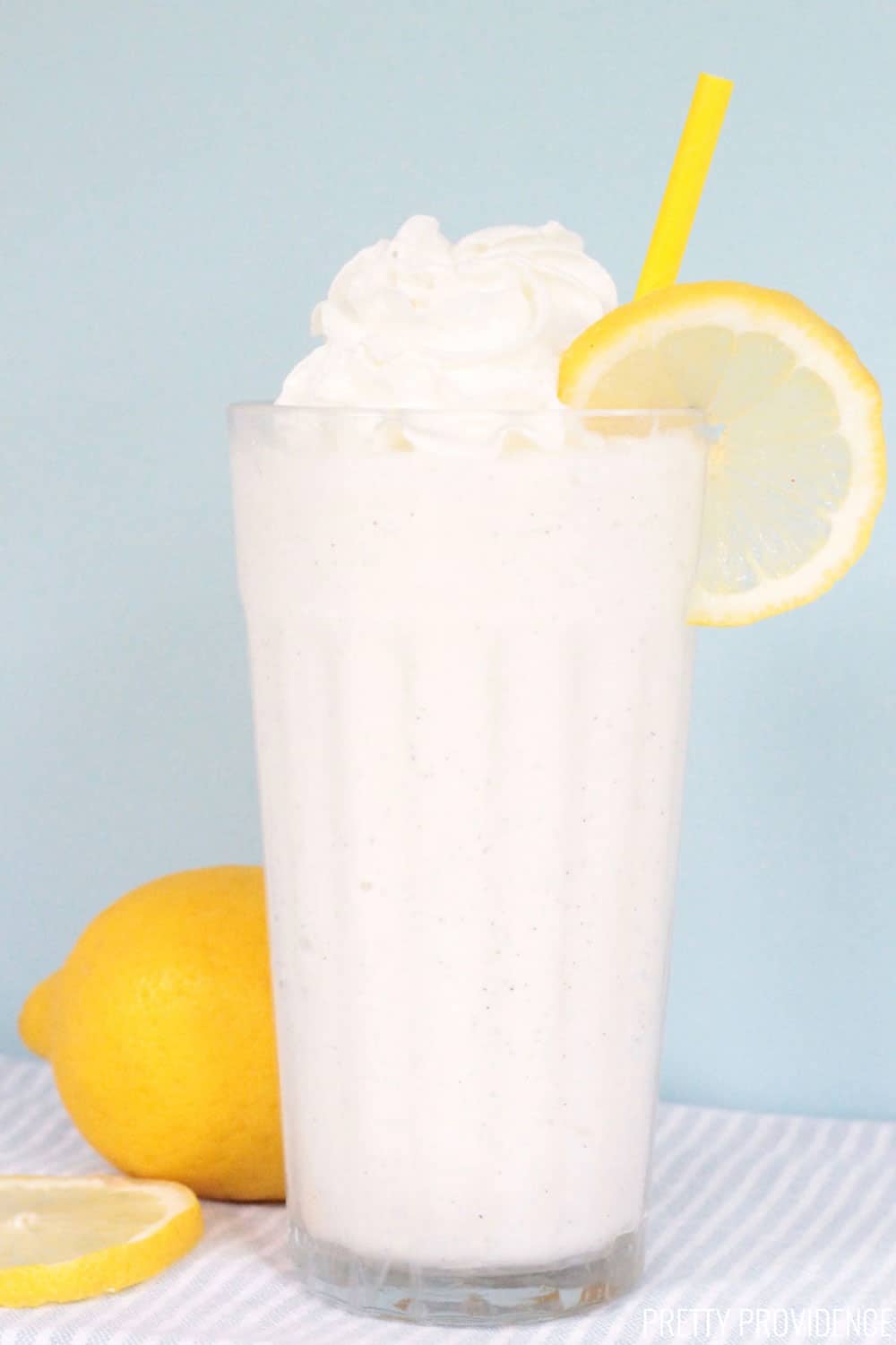 Frosted Lemonade in a tall glass with a yellow straw, topped with whipped cream and a lemon slice.