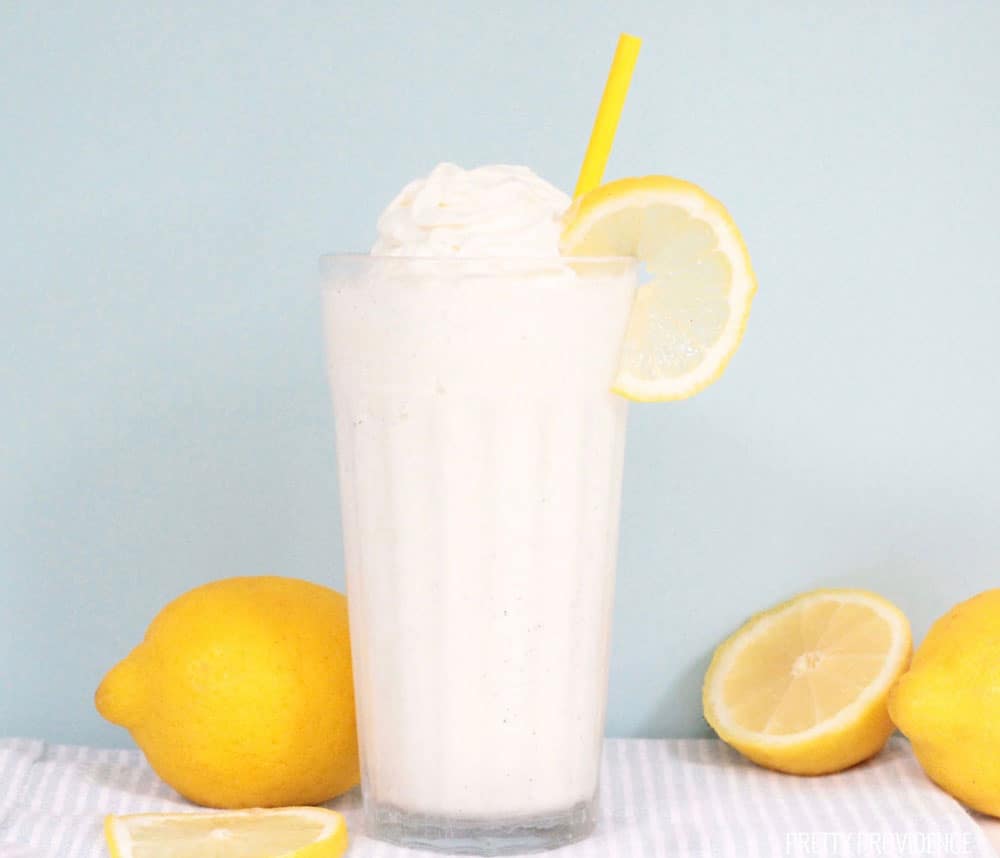 Frosted Lemonade in a glass topped with whipped cream and a lemon slice and surrounded by fresh lemons.
