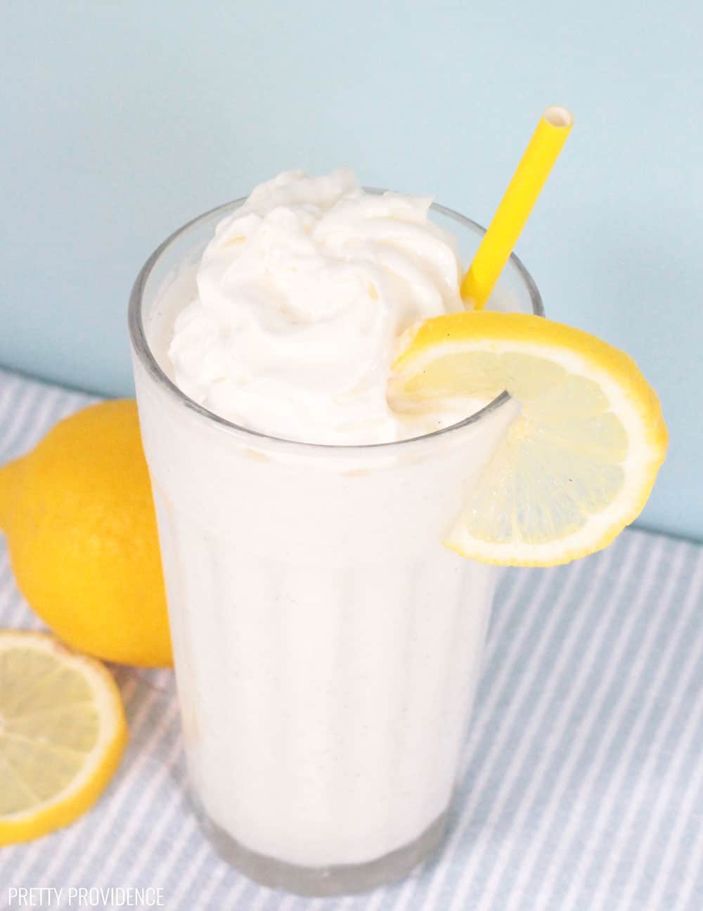 Frozen lemonade in a clear glass with ice cream, whipped cream on top, and garnished with a lemon slice and yellow straw.