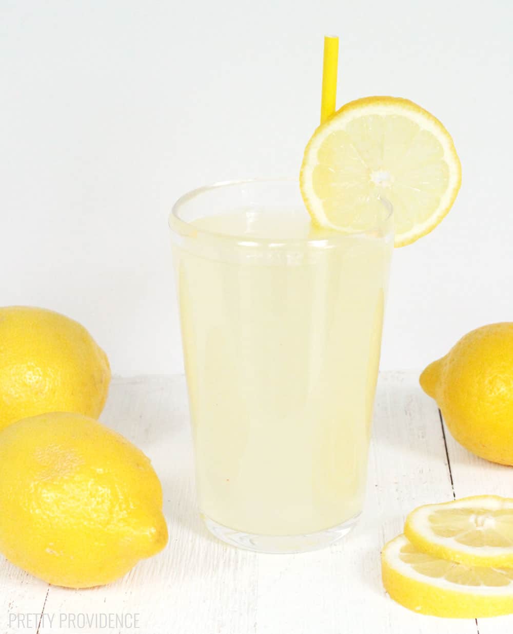 Fresh squeezed lemonade in a clear glass with lemon slice and yellow straw as garnish and lemons around it on the sides.