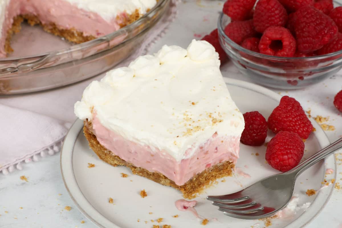 A slice of raspberry cream cheese pie with graham cracker crust topped with homemade whipped cream, a fork resting next to it on a white plate.