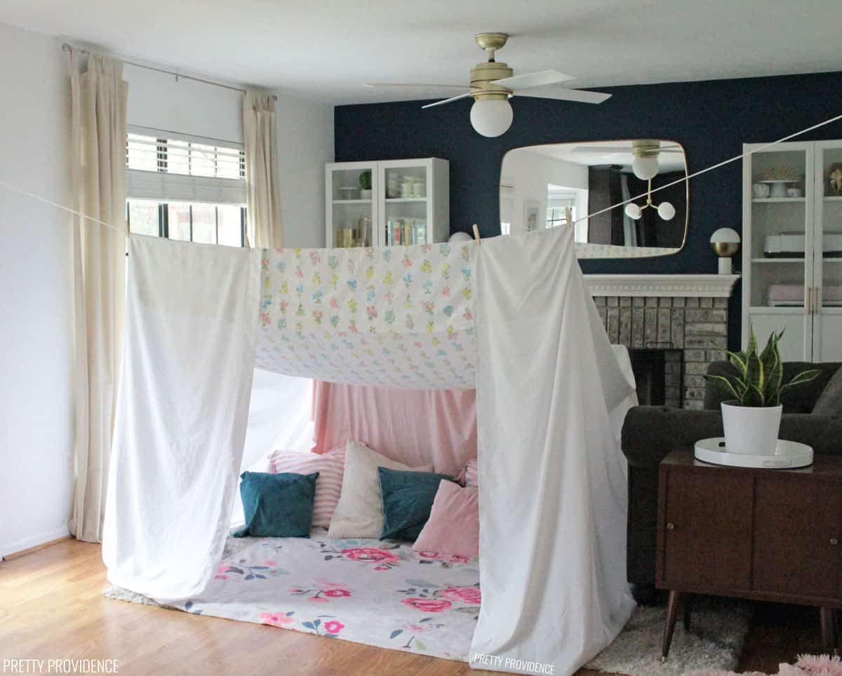 Blanket fort in a living room made with bedsheets, clothes pins, and clothesline tied from door hinge to door hinge. Inside the fort are pink, blue and cream colored pillows.