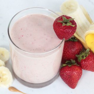 Strawberry banana protein smoothie in a glass with strawberries, a peeled banana and sliced banana surrounding the glass.