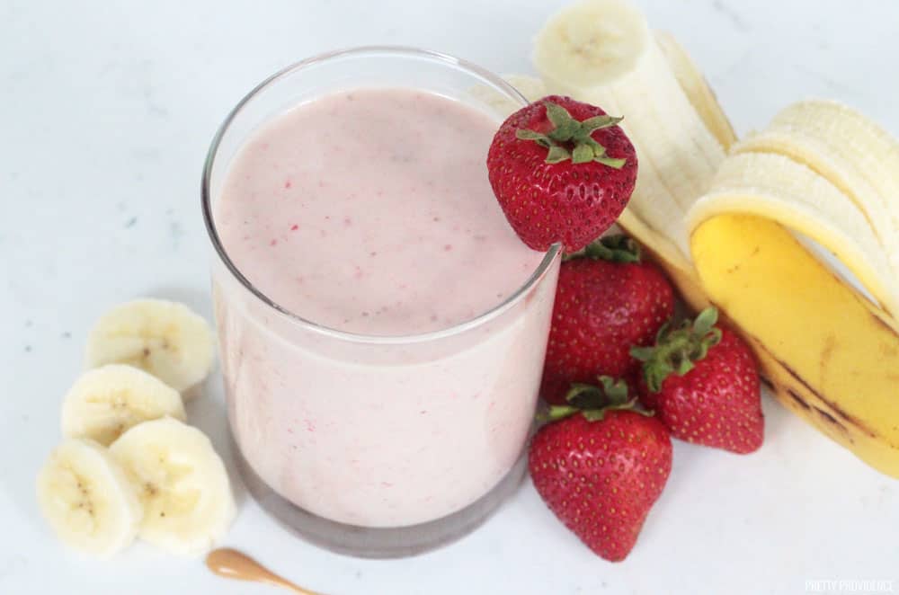 Strawberry banana protein smoothie in a glass with strawberries, a peeled banana and sliced banana surrounding the glass.