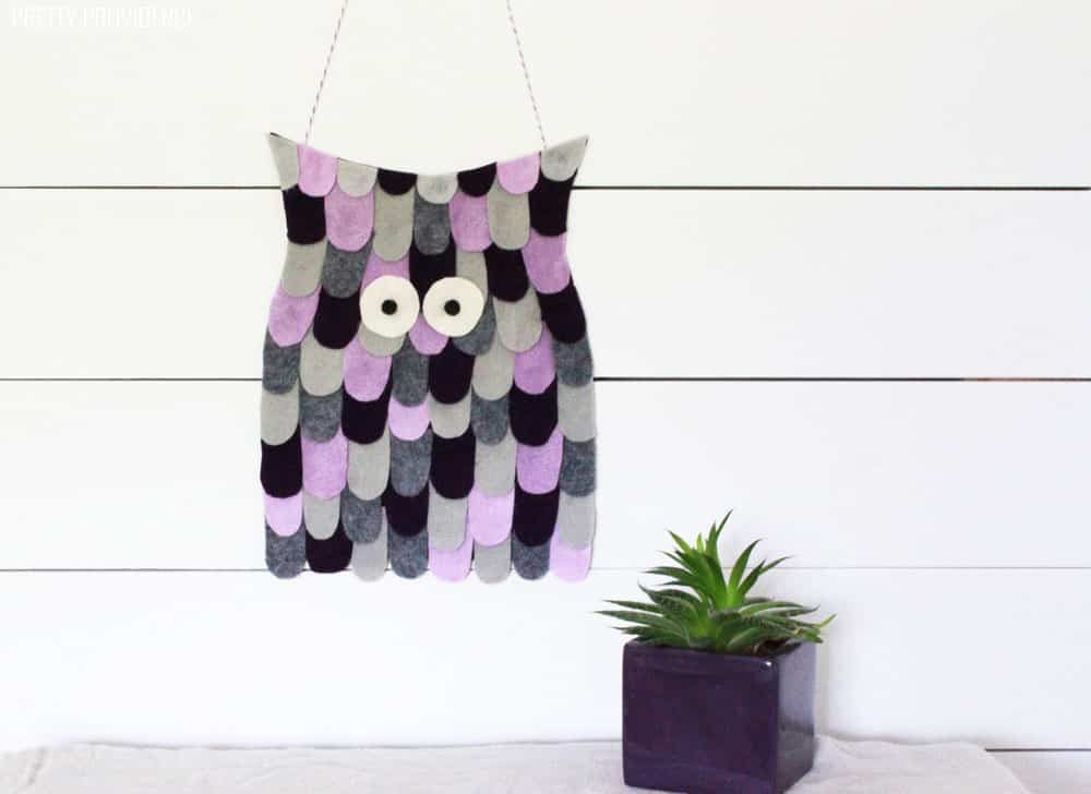 Felt owl Halloween decoration on a white wall with a succulent plant next to it.