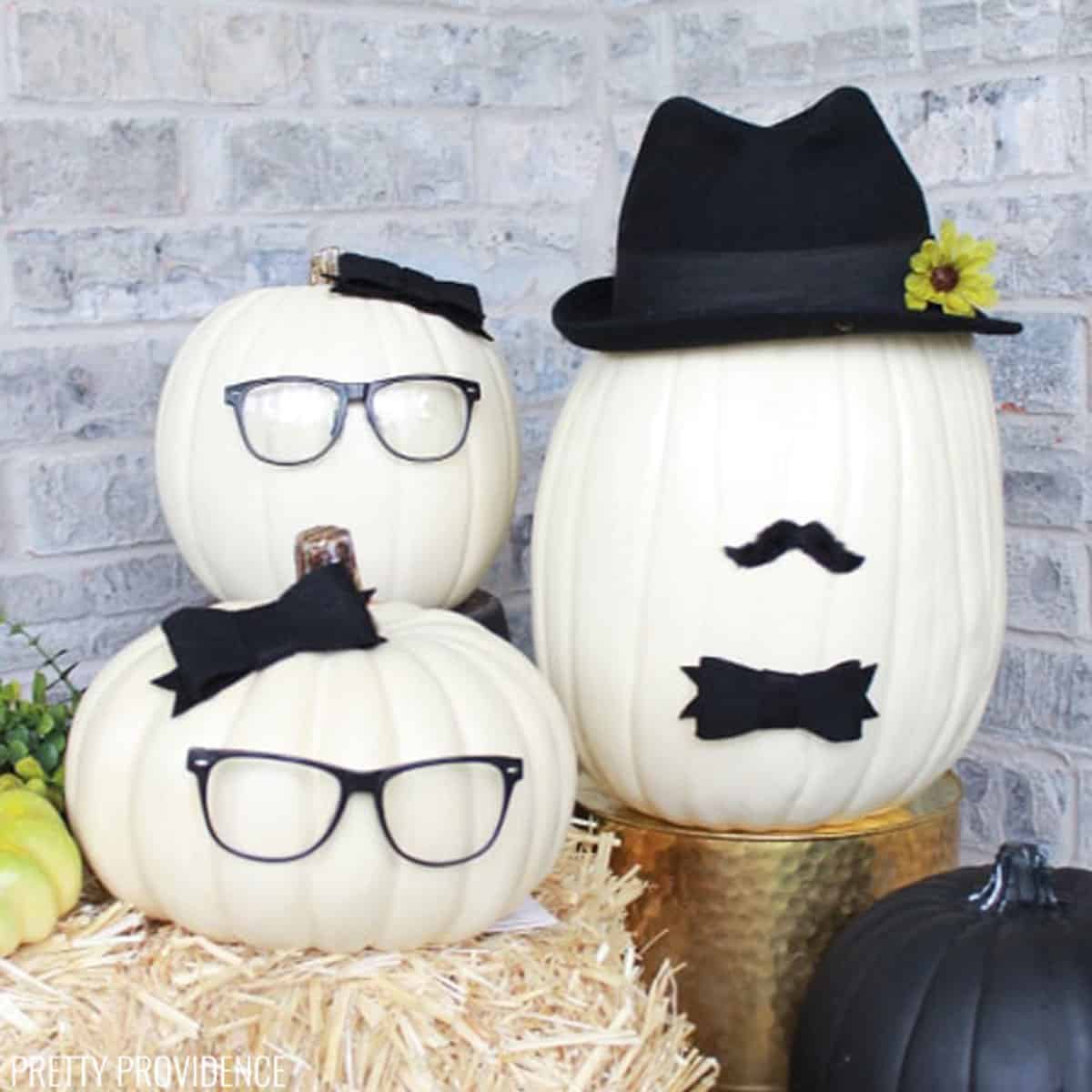 Three cream-colored pumpkins. Two pumpkins have black eyeglasses and bows glued on, and one has a top hat, a mustache and bow tie glued on.