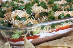 Classic Seven Layer Dip in a glass dish topped with cheese and green onions with tortilla chips
