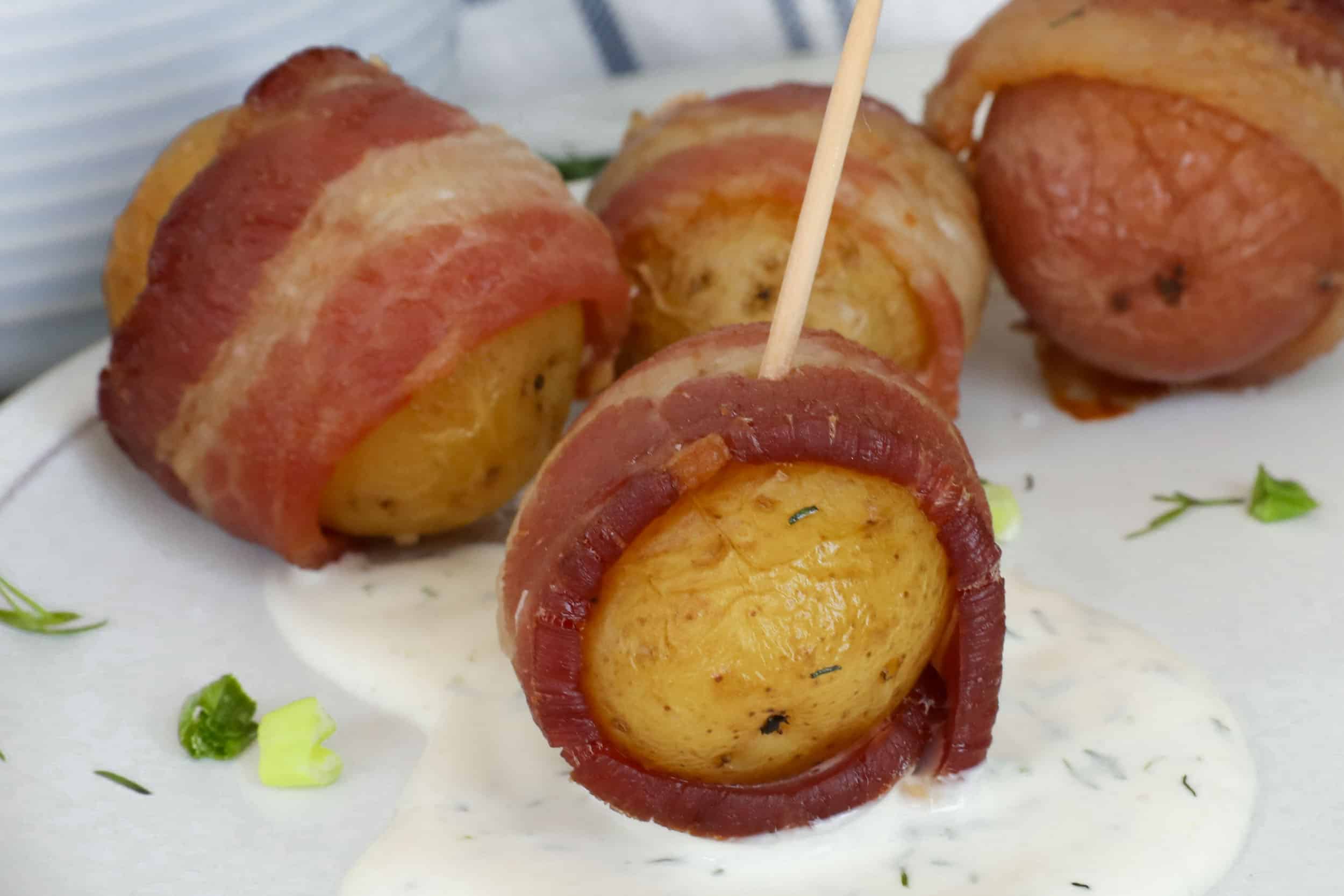 Bacon Wrapped Potatoes with toothpicks on a white plate with creamy dill sauce on the side.