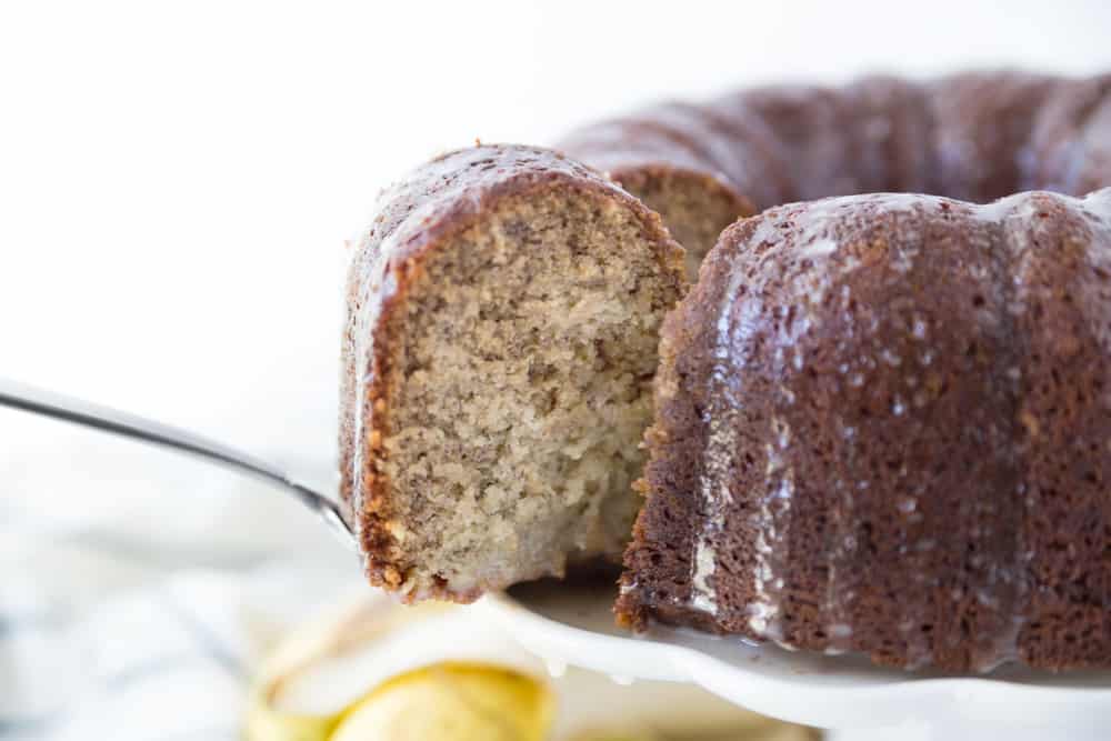 Banana cake on a white cake plate with one slice being removed from it.