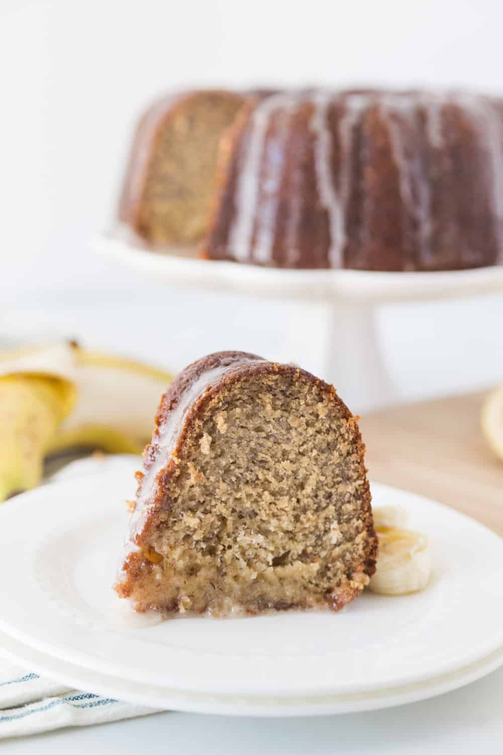 Slice of banana cake, garnished with banana slices and with the rest of the cake on a white cake plate behind it.