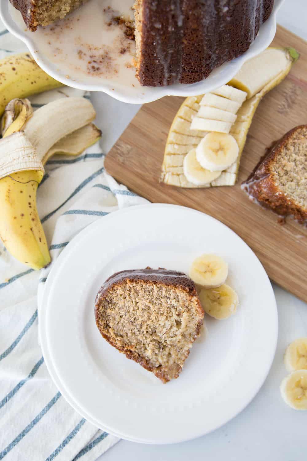 Banana cake on a white plate with bananas sliced as garnish and a beautiful cutting board under it.