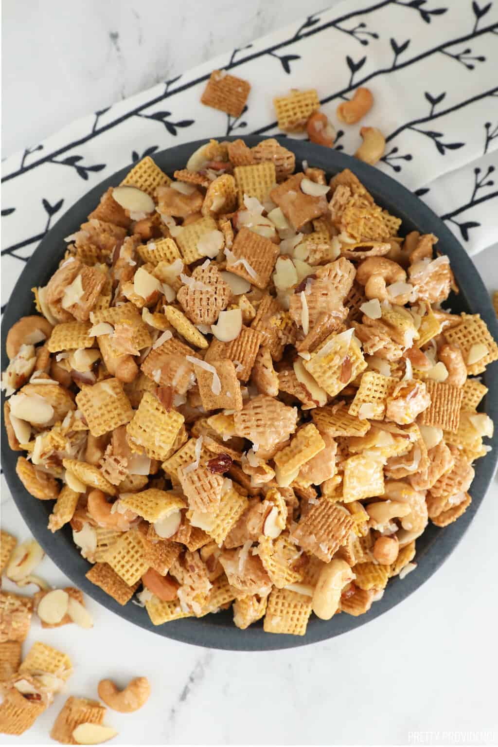 Gooey Chex mix - close up of Chex, cashews, coconut, almonds with sticky caramel coating in a white bowl.