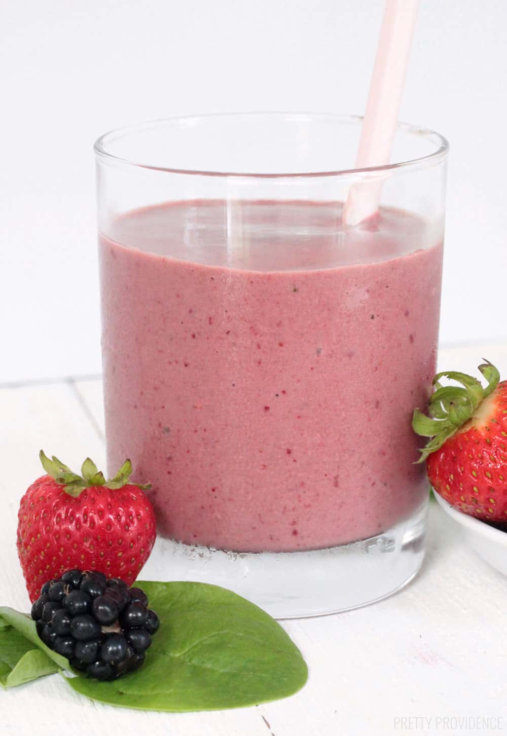 Berry Spinach Smoothie in glass, a pink straw, and strawberries, spinach and blackberries surrounding it.