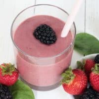 Berry Spinach Smoothie in a glass, topped with a fresh blackberry, and spinach leaves, strawberries and blackberries surrounding the glass.