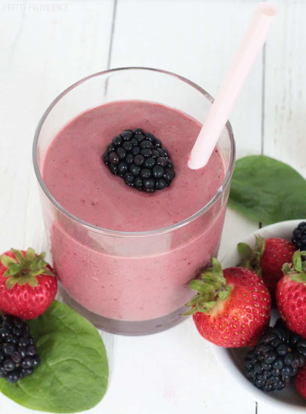 Berry Spinach Smoothie in a glass, topped with a fresh blackberry, and spinach leaves, strawberries and blackberries surrounding the glass.