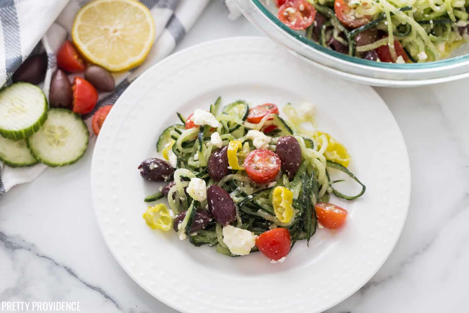 Spiralized zucchini recipe for authentic greek salad on a white plate with kalamata olives, pepperoncini, tomatoes and feta cheese.