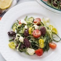 Greek salad on a white plate, spiralized zucchini, tomatoes, kalamata olives, pepperoncini, feta cheese and greek salad dressing.