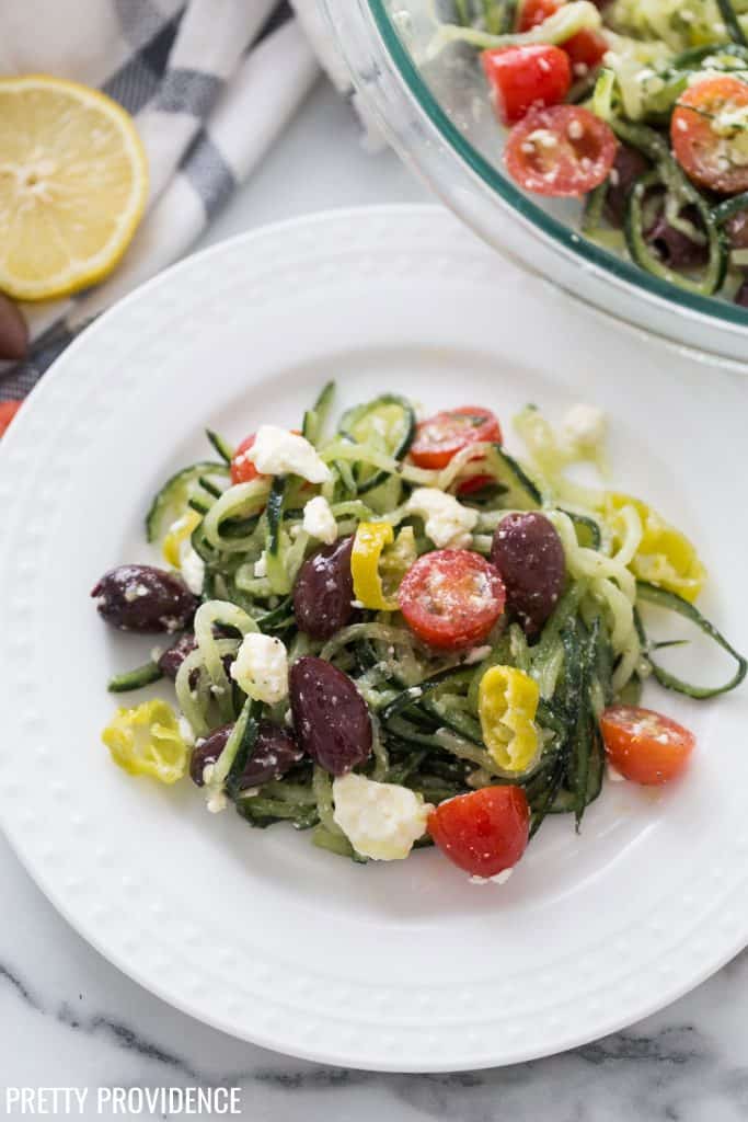 Greek salad on a white plate, spiralized zucchini, tomatoes, kalamata olives, pepperoncini, feta cheese and greek salad dressing.
