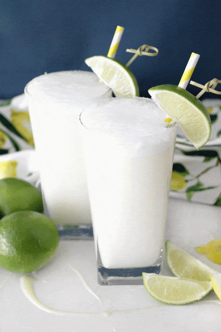 Two glasses of brazilian lemonade garnished with limes against a blue wall