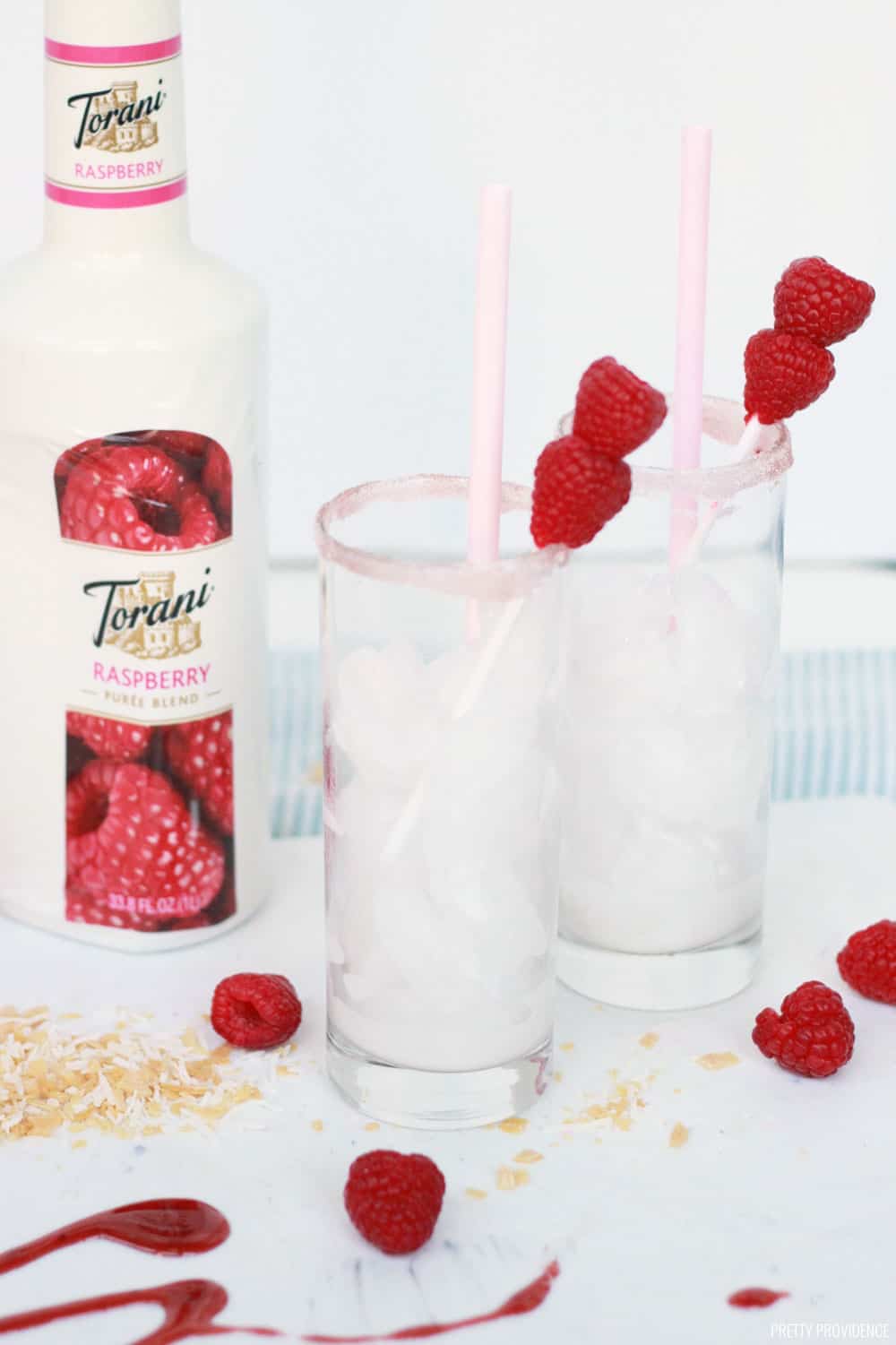 Two highball glasses filled with ice, garnished with fresh raspberries and Torani Raspberry puree next to it on the table.