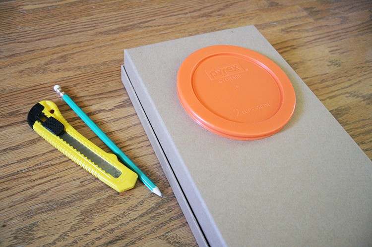 Brown cardboard box with a pyrex lid on it, an exacto knife and a pencil beside them.