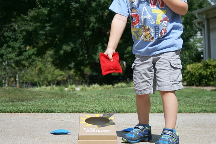 A small child holding a bean bag next to a mini corn hole board.