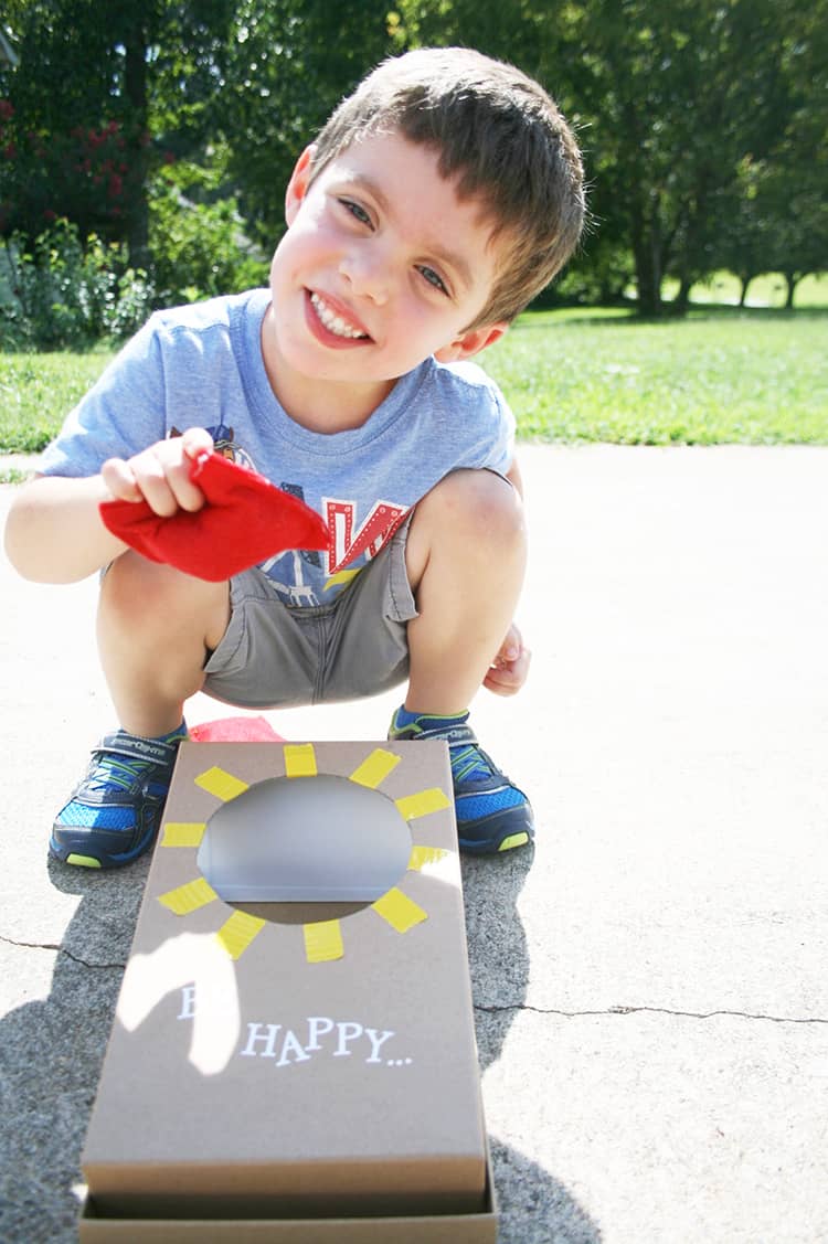 A small boy holding a red bean bag and smiling by a mini corn hole board.