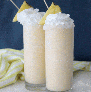 two tall glasses filled with pina colada slushes garnished with pineapple against a blue background
