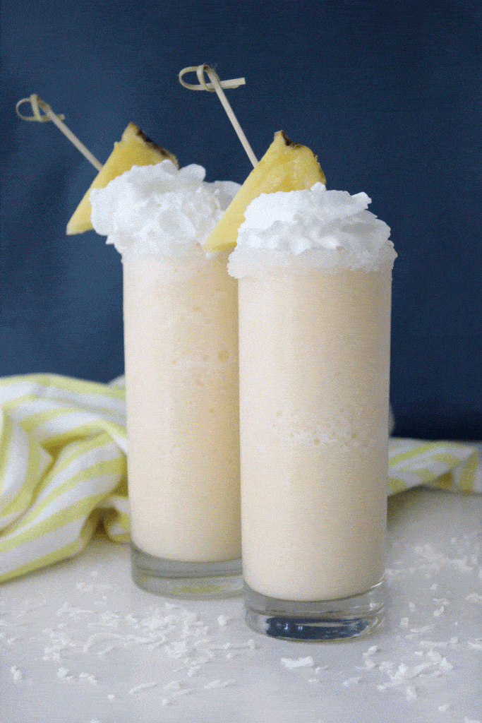 two tall glasses filled with pina colada slushes garnished with pineapple against a blue background